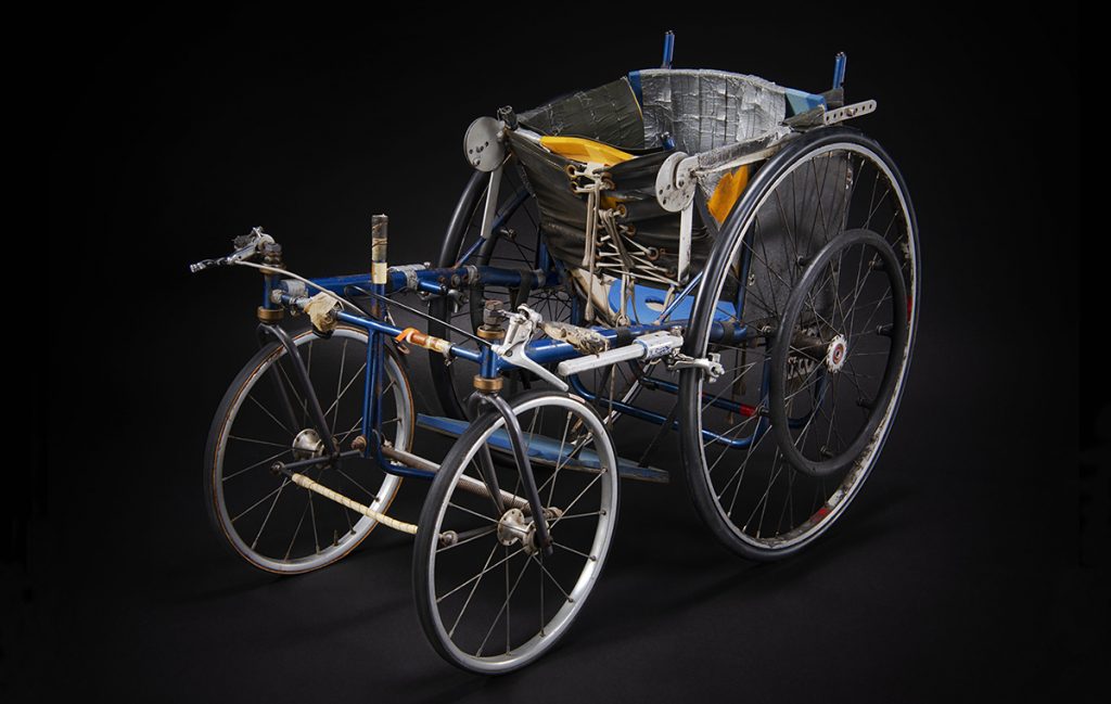 Un fauteuil roulant comprenant une armature métallique bleue, quatre roues et un siège baquet recouvert de ruban adhésif argenté