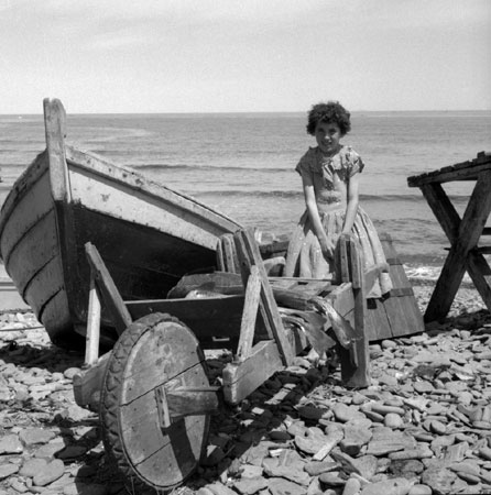 Girl sitting on a barrel next to a fishing boat on the shore, [19--]., © CMC/MCC, J15267