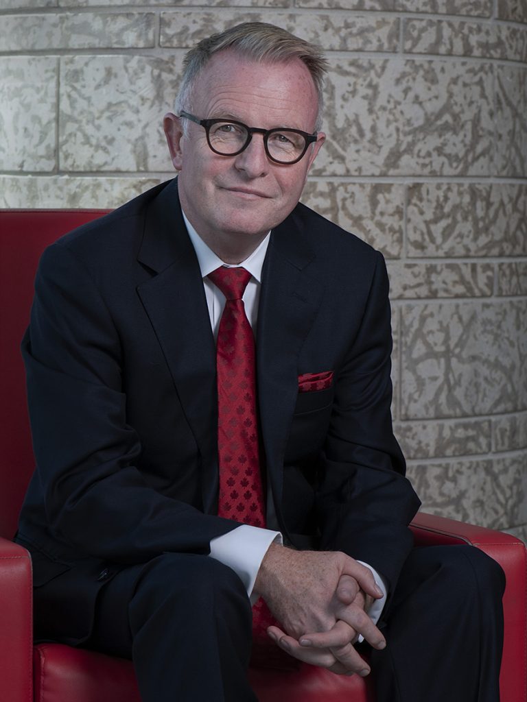Portrait d’un homme souriant, assis dans un fauteuil rouge et portant un complet foncé, une chemise blanche, une cravate rouge et des lunettes