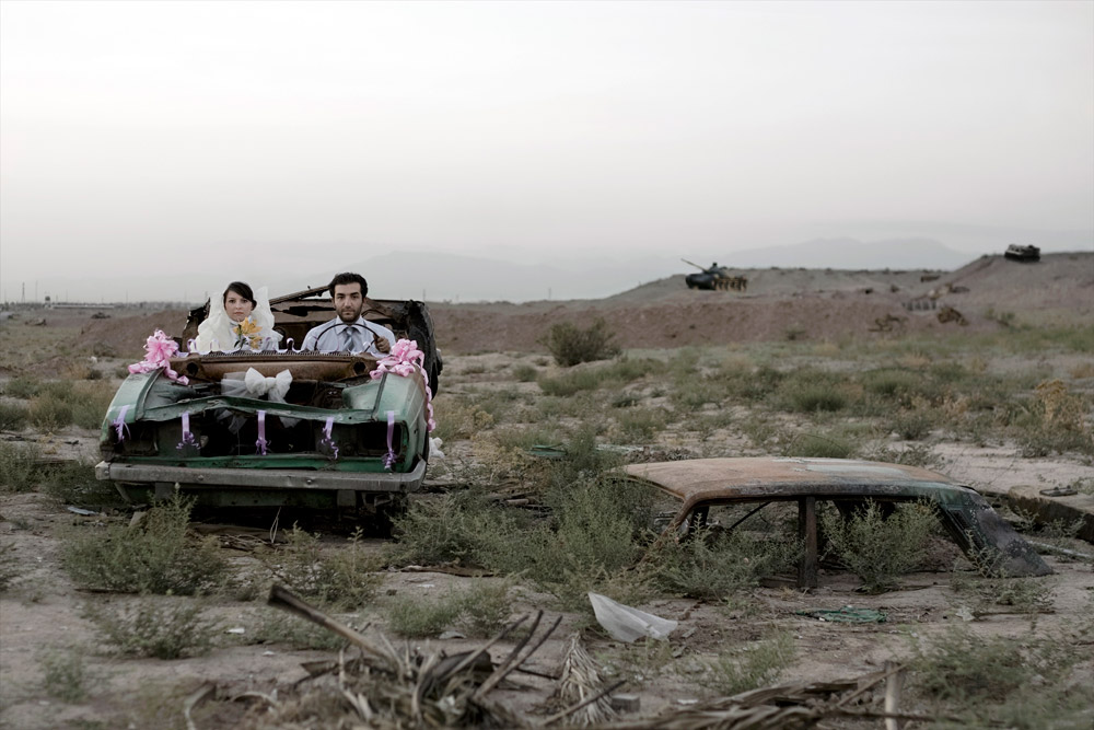 Un couple nouvellement marié prenant place dans une vieille voiture devant un paysage ravagé par la guerre