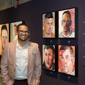 Un homme souriant à côté d’un mur couvert de portraits, dont un le représente
