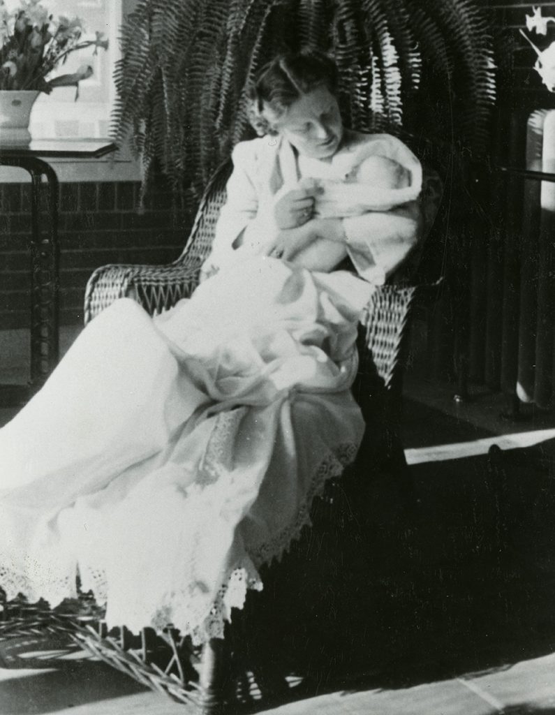 Photo en noir et blanc d’une femme vêtue d’une longue robe blanche et tenant un bébé, assise dans un fauteuil en osier tressé