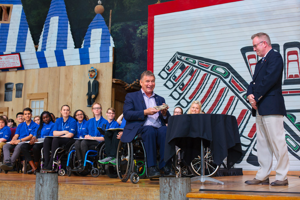 Hansen, dans un fauteuil roulant, tenant son gant sur la scène de la Grande Galerie en compagnie d’étudiantes et d’étudiants ainsi que de Mark O’Neill