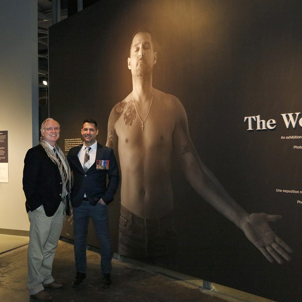 Deux hommes posent devant une grande photographie d’un homme au torse nu avec des tatouages et une longue cicatrice au bras