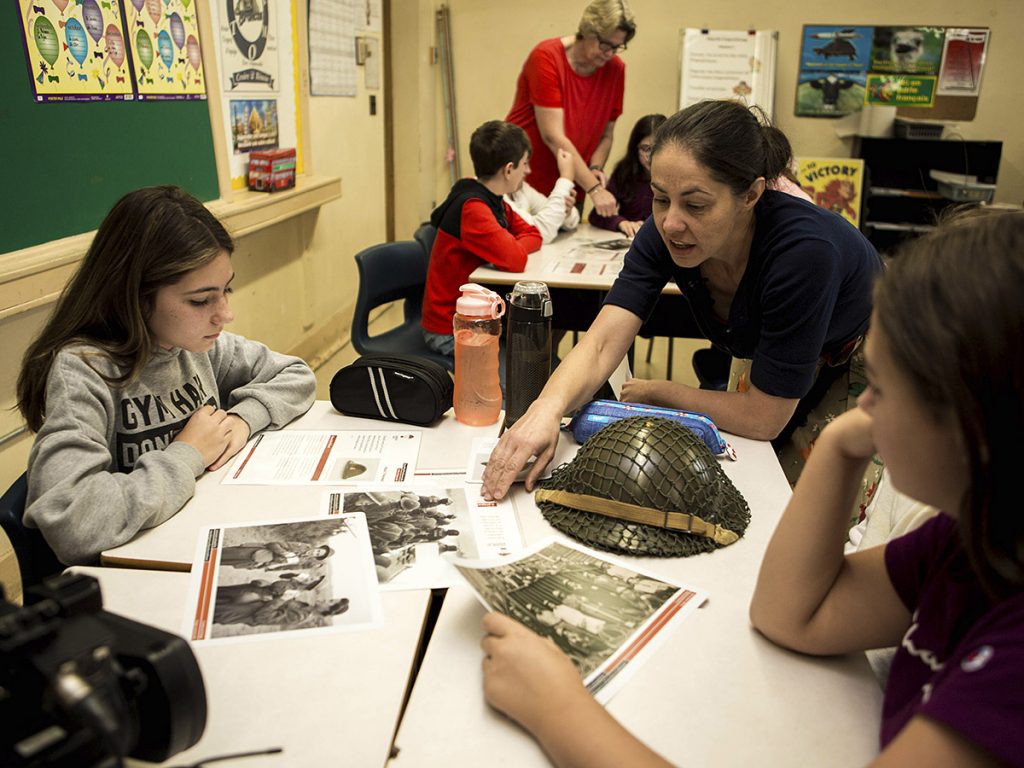 Une femme parle à deux adolescentes assises en classe autour d’une table sur laquelle se trouvent un casque militaire et des dépliants