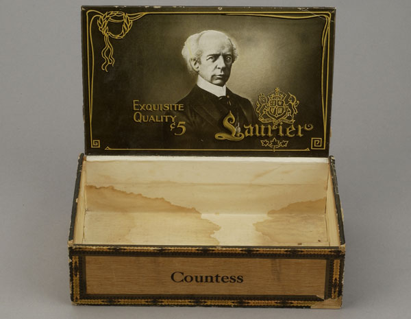 Ancienne boîte à cigares en bois rectangulaire avec couvercle à charnière. Le nom « Laurier » dans une couronne de laurier est gravé sur le dessus du couvercle. L’intérieur est orné d’une photographie noir et blanc de Laurier et d’un écusson doré. Musée canadien de l’histoire, 1999.124.21