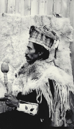 Ususllst, a Nlaka'pamux (Thompson) man wearing traditional clothing (fringed fur cape, woven bark and fur hat, beaded cuffs and rattle) , profile view, photographed in 1913 at Spences Bridge, British Columbia, © CMC/MCC, J.A. Teit, 20831