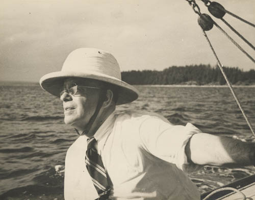 Un homme à la barre d’un voilier portant un casque colonial, des lunettes et une cravate, avec de l’eau et des arbres en arrière-plan.