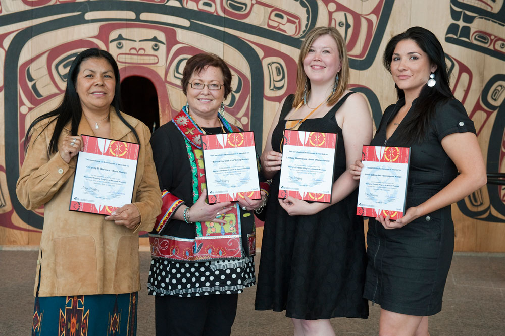 Quatre femmes détenant des certificats devant un totem au Musée canadien de l'histoire à Ottawa.