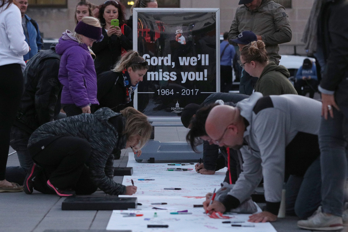Plusieurs adultes s’agenouillent sur le sol, écrivant sur une grande feuille de papier avec des marqueurs. D’autres adultes et enfants se tiennent à proximité et un grand panneau en arrière-plan indique « Gord, we’ll miss you! 1964-2017. » (Gord, tu vas nous manquer! 1964-2017).