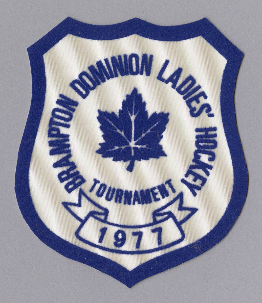 Un insigne bleu et blanc sur lequel figure une feuille d’érable entourée des mots « Brampton Dominion Ladies’ Hockey Tournament », sous lesquels se trouve une bannière sur laquelle est inscrit le chiffre « 1977 »