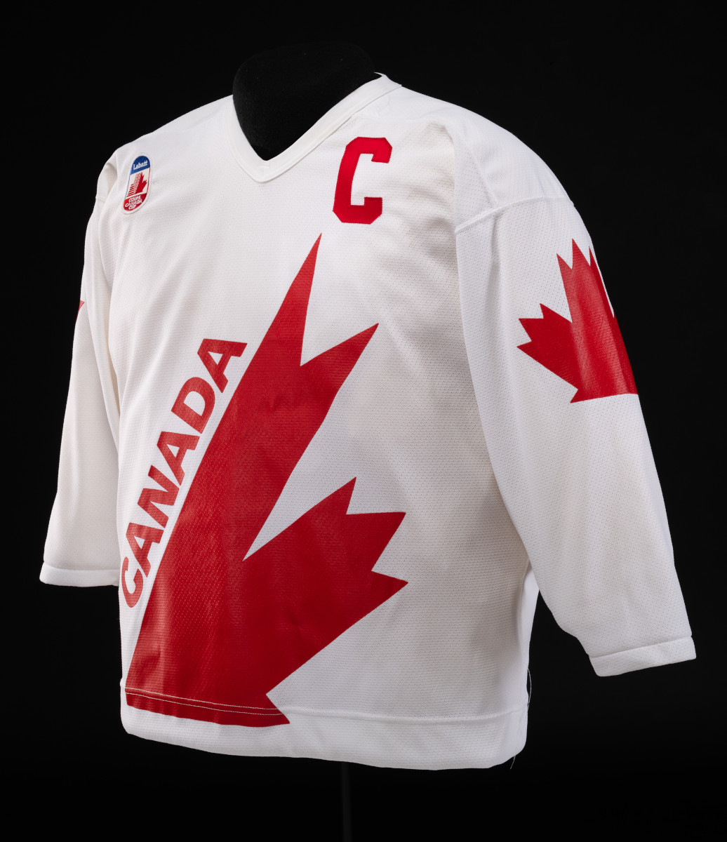 Un maillot de hockey blanc sur lequel figurent une grande feuille d’érable rouge en diagonale et le mot « CANADA » sur le devant, un grand « C » sur la partie avant de l’épaule gauche et une feuille d’érable plus petite sur la manche.
