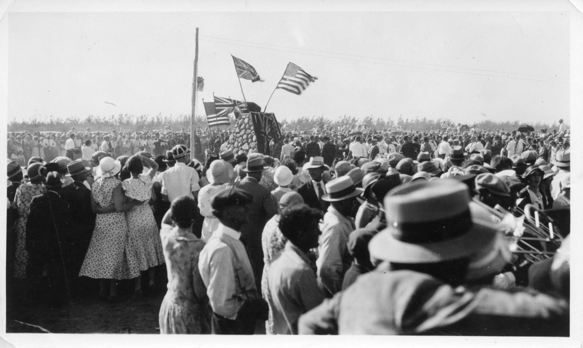 Photo en noir et blanc d’une grande foule entourant un cairn de pierre arborant l’Union Jack et le drapeau des États-Unis.