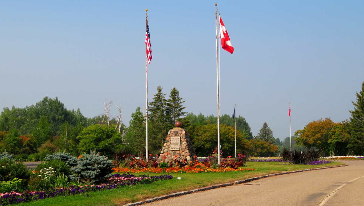 Un cairn de pierre avec une plaque sculptée se dresse entre deux mâts, l’un arborant le drapeau du Canada, l’autre, le drapeau des États-Unis.