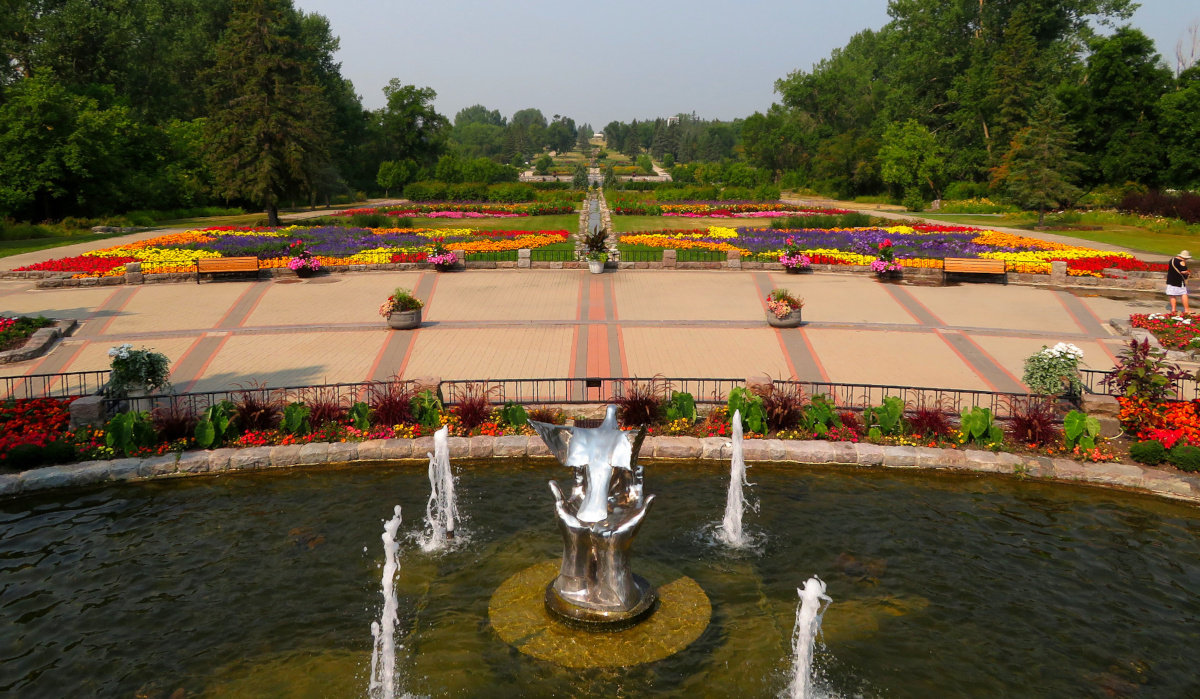 Un vaste jardin de fleurs aménagé avec un bassin d’eau et une fontaine au premier plan. Au centre de la piscine se trouve une sculpture en métal brillant représentant une paire de mains libérant une colombe.