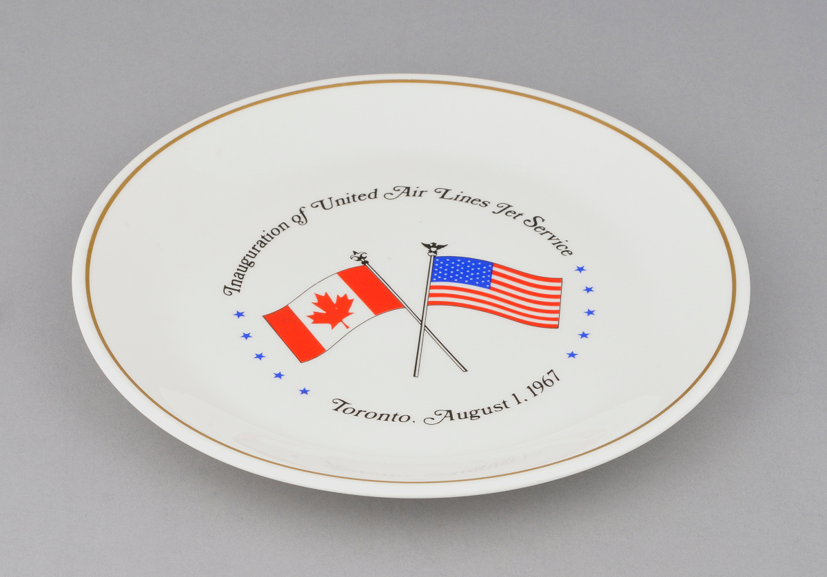 Une assiette en céramique blanche illustrant deux drapeaux croisés, celui du Canada et celui des États-Unis, entourés de texte disant « Integration of United Airlines for Service, Toronto, August 1, 1967 » (Intégration du service d’United AirLines, Toronto, 1er aout 1967).