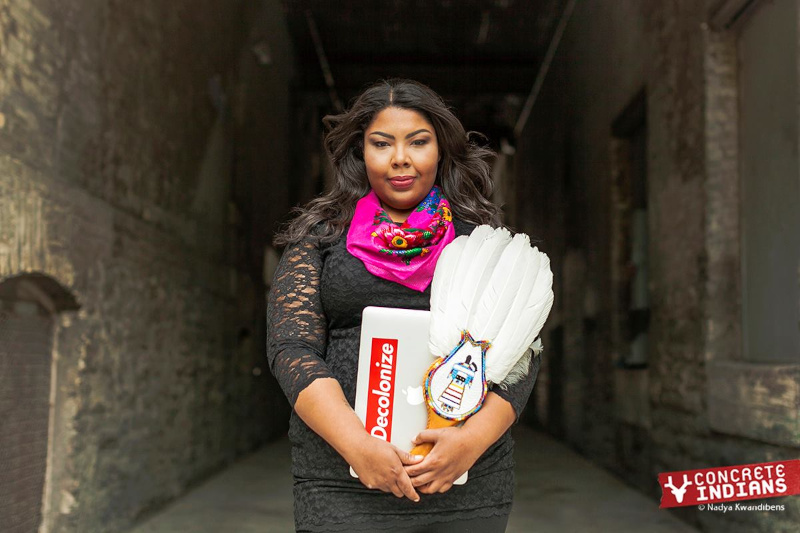 Une Autochtone aux cheveux longs porte une robe en dentelle noire et un foulard floral rose, et tient un éventail de cérémonie à plumes blanches orné de perles et un ordinateur portable avec un autocollant « Decolonize » (Décoloniser).