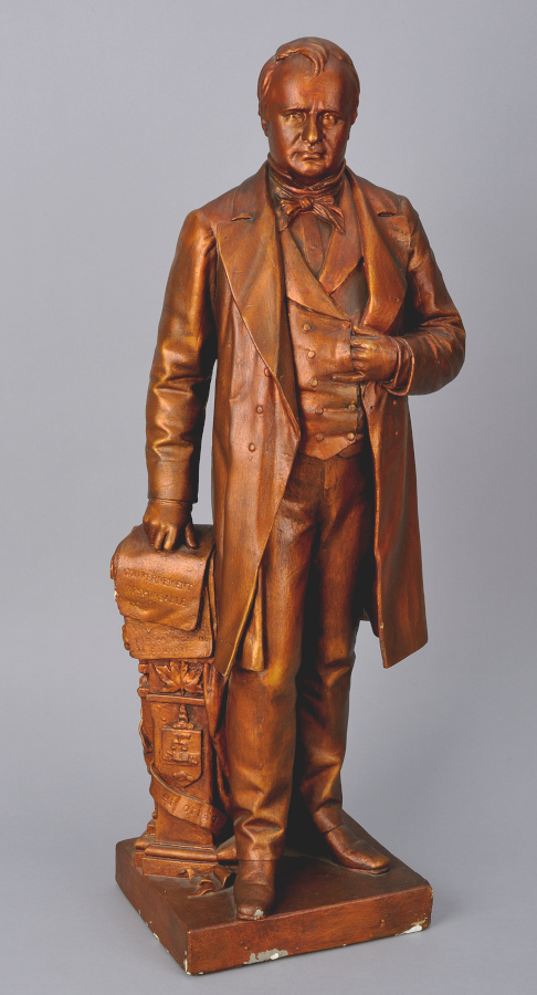 Une statue en bois représentant un homme en tenue de cérémonie, appuyé d'une main sur une pile de livres et l'autre main glissée dans son gilet.