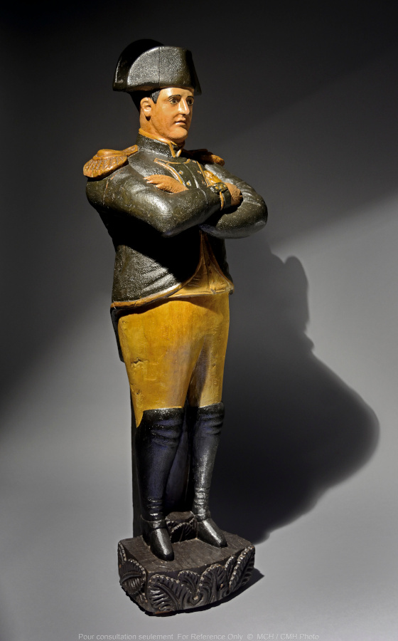 Une statue représentant un homme coiffé d'un grand chapeau et chaussé de bottes hautes, debout, les bras croisés.