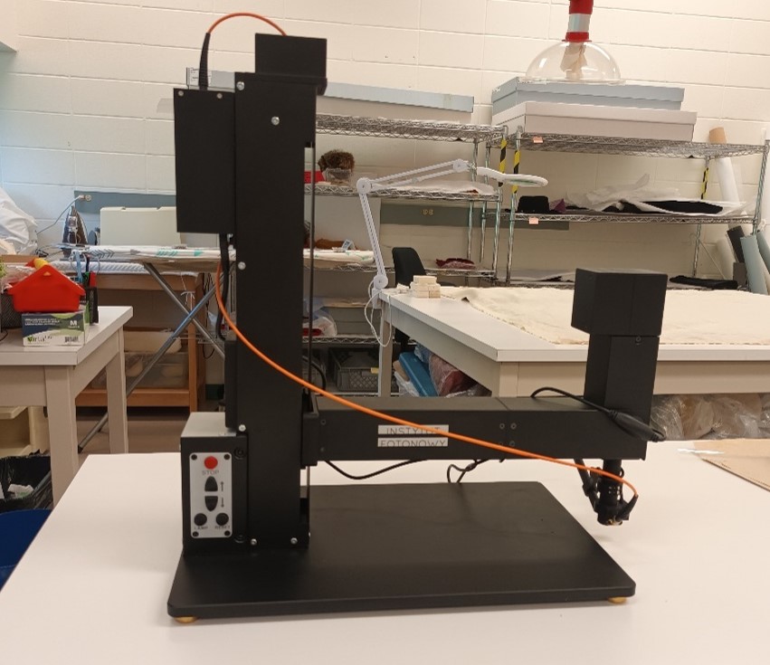 Une machine noire avec une plaque de base plate et une colonne verticale carrée, d’environ un demi-mètre de haut et de large. Un bras horizontal part de la colonne et est légèrement plus long que la base.