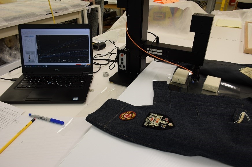 Une veste sombre ornée d’insignes militaires placée sous le bras saillant d’un appareil noir relié à un ordinateur portable.